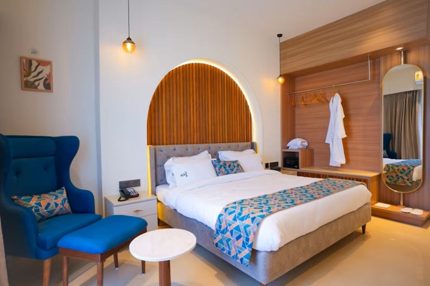 A hotel room with a large bed, a blue armchair, a small table, a bedside table with a lamp, and a mirror.