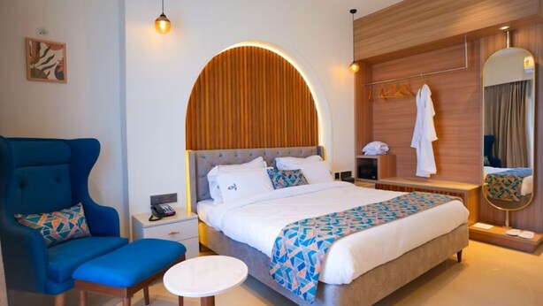 A hotel room with a large bed, a blue armchair, a small table, a bedside table with a lamp, and a mirror. - THE ELET (Amritsar)