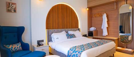 Habitación de hotel con una cama grande, un sillón azul, una mesita, una mesita de noche con lámpara y un espejo.