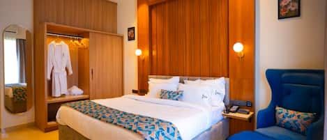 A hotel room with a large bed, a seating area with a blue chair, a wardrobe, and a mirror.