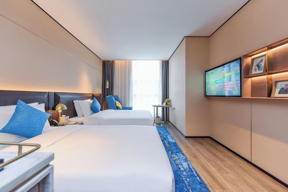 Ein Hotelzimmer mit zwei Betten, einem Fernseher an der Wand, einem kleinen Tisch und einem blauen Teppich.