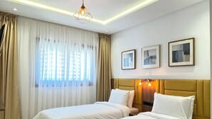 Une chambre d’hôtel avec deux lits, une grande fenêtre avec des rideaux, un luminaire au plafond et des tableaux encadrés accrochés au mur.