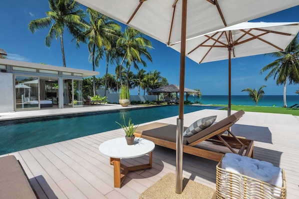 A poolside area with a lounge chair, a table, and a view of the ocean and palm trees.