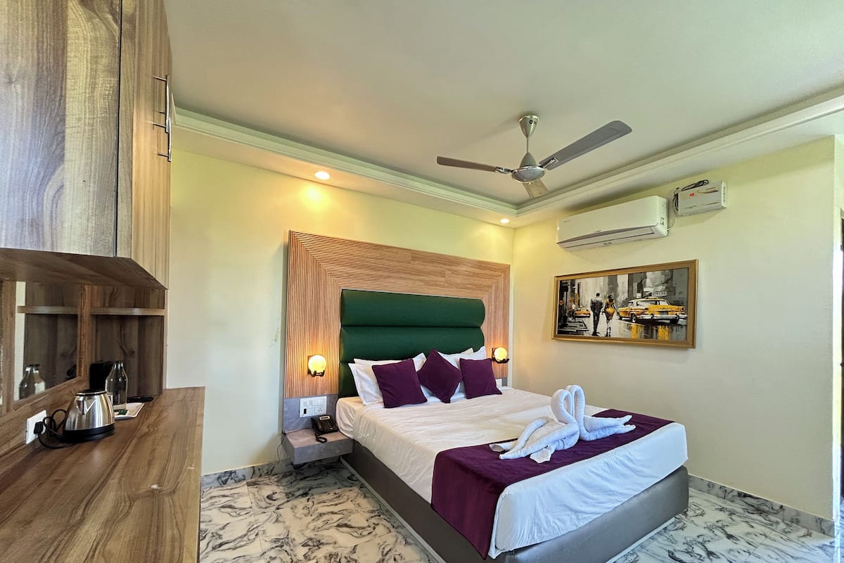 A hotel room with a bed, a desk, a ceiling fan, an air conditioning unit, and a painting on the wall.