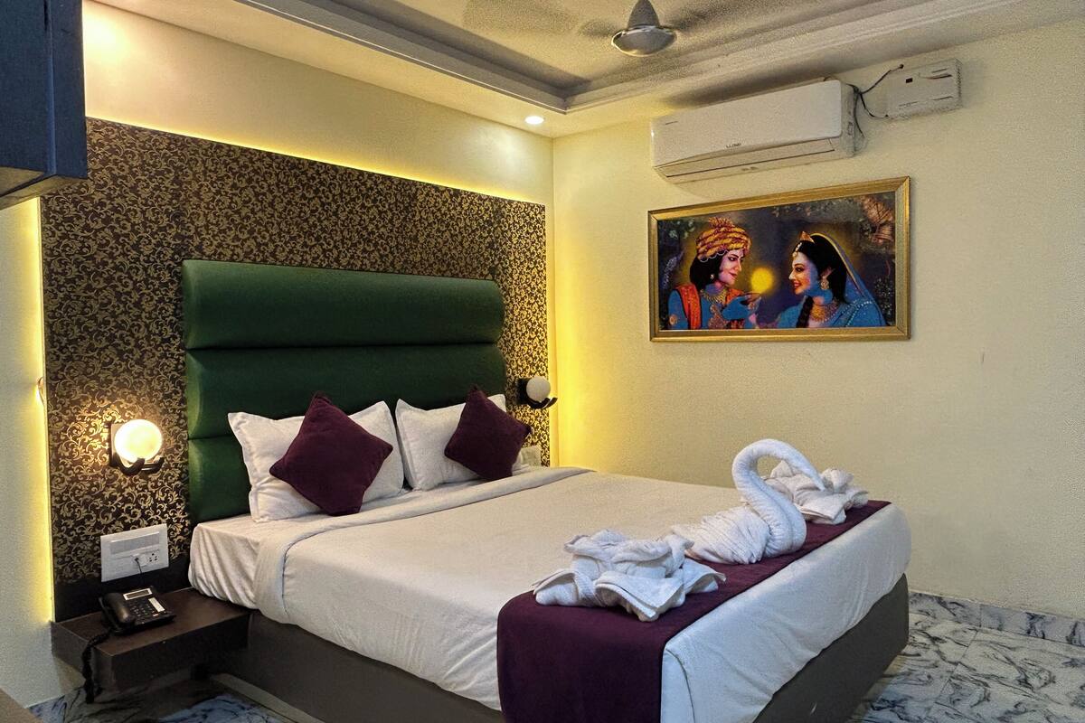 A hotel room with a green upholstered headboard, a bed with white and purple pillows, a painting on the wall, and a towel on the bed.