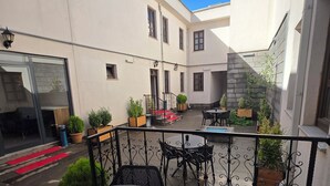 Μια αυλή με ένα στρογγυλό τραπέζι και καρέκλες, γλάστρες με φυτά και ένα κόκκινο χαλί που οδηγεί σε μια πόρτα.