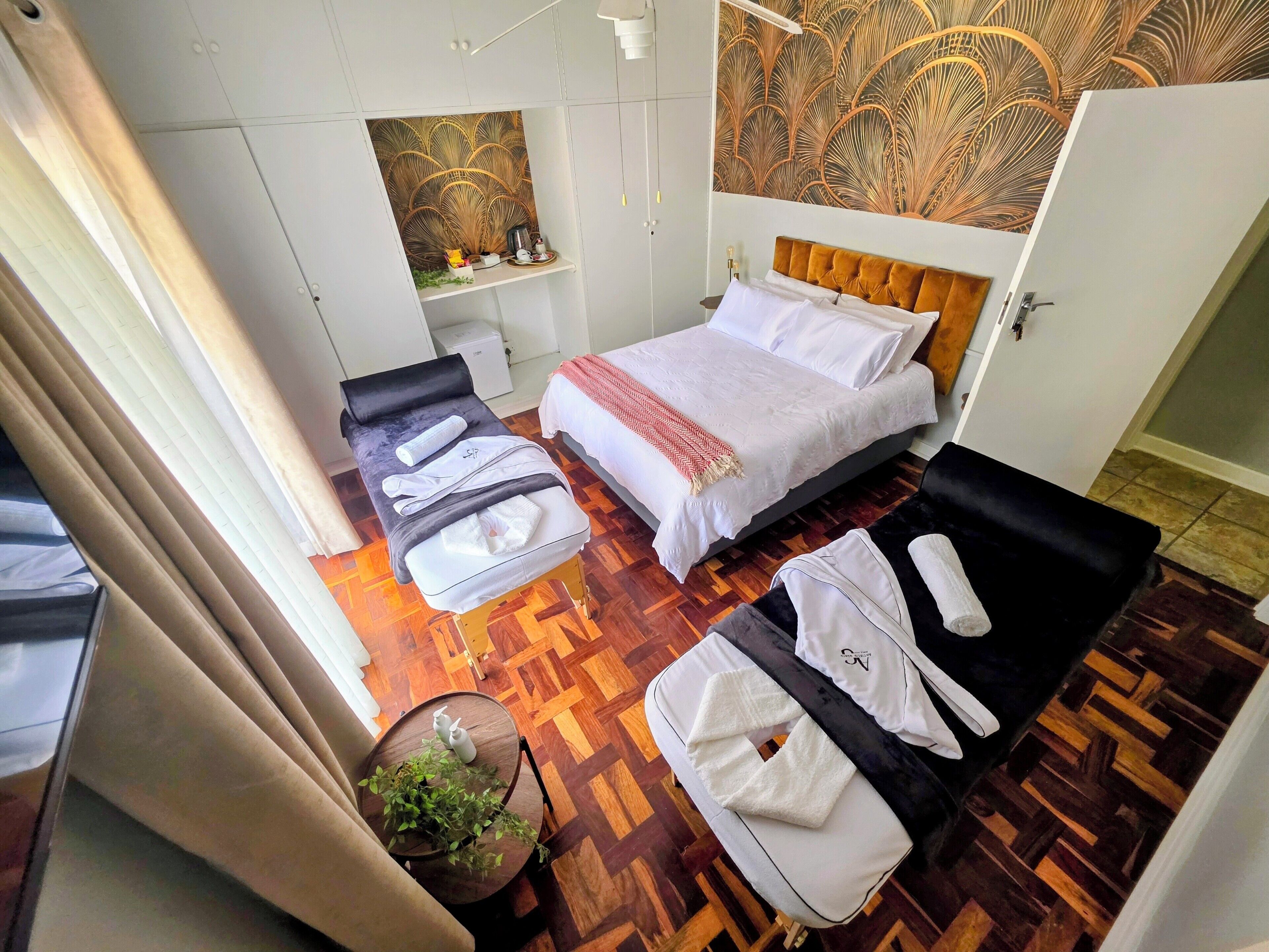 A hotel room with a bed, a massage table, a small table with a plant, and a wall with a decorative pattern.