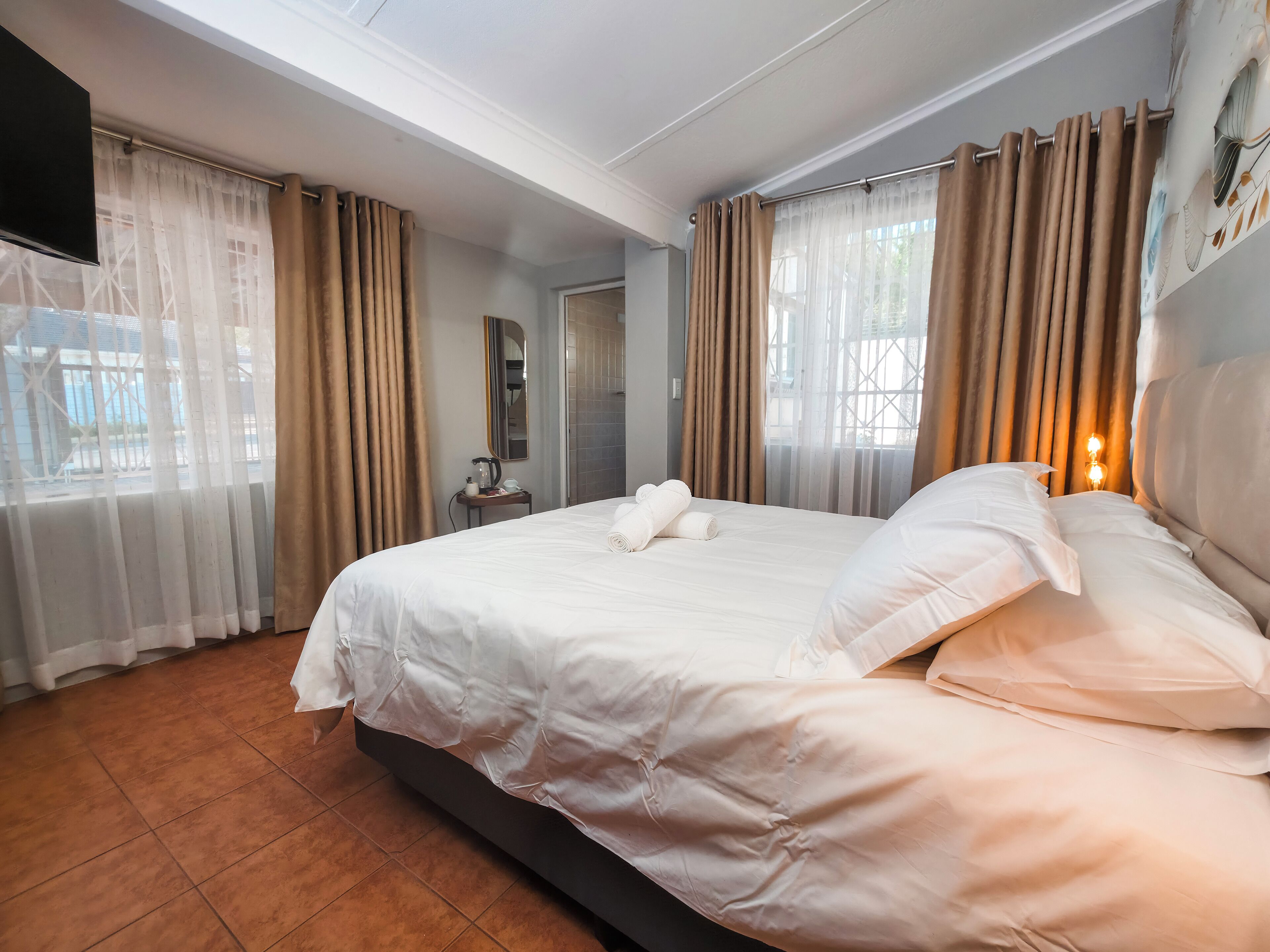 A hotel room with a large bed, a bedside table with a lamp, and a view of the outside through sheer curtains.