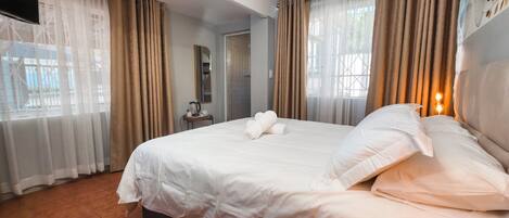 A hotel room with a large bed, a bedside table with a lamp, and a view of the outside through sheer curtains.