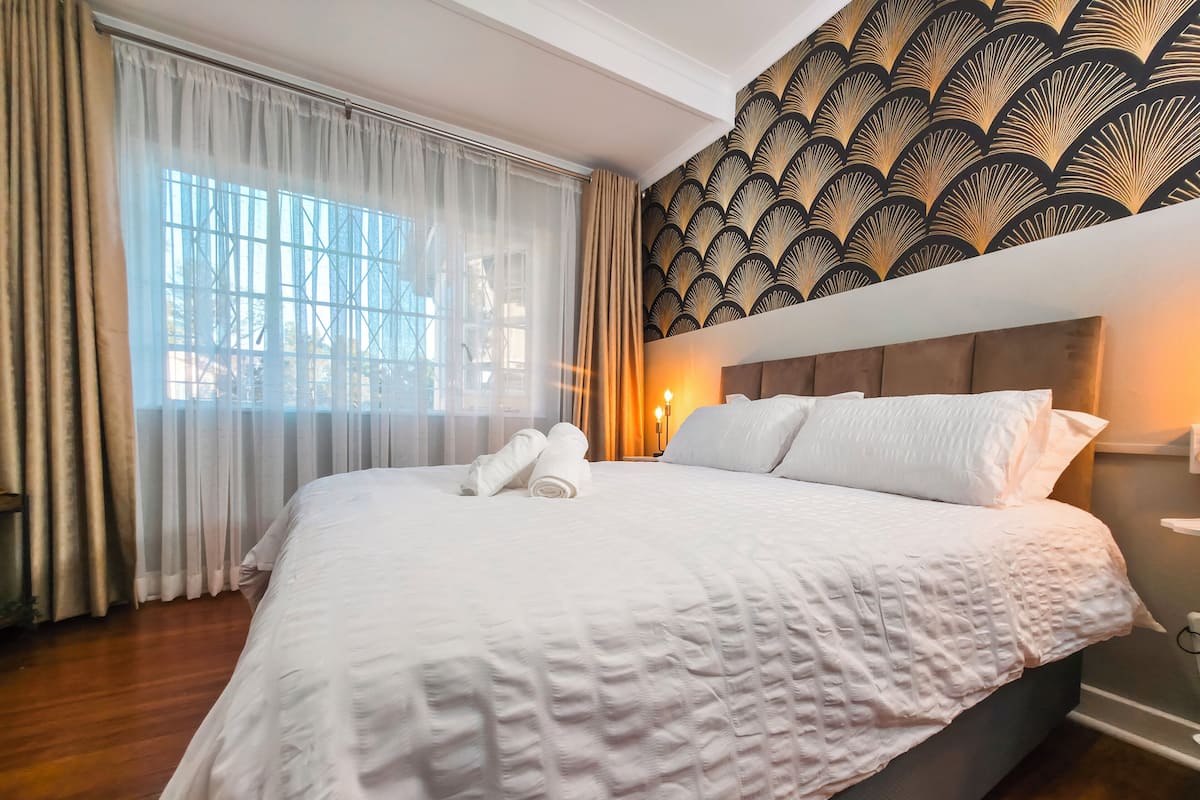 A neatly made bed with white linens, a headboard with a patterned design, and a window with sheer curtains.
