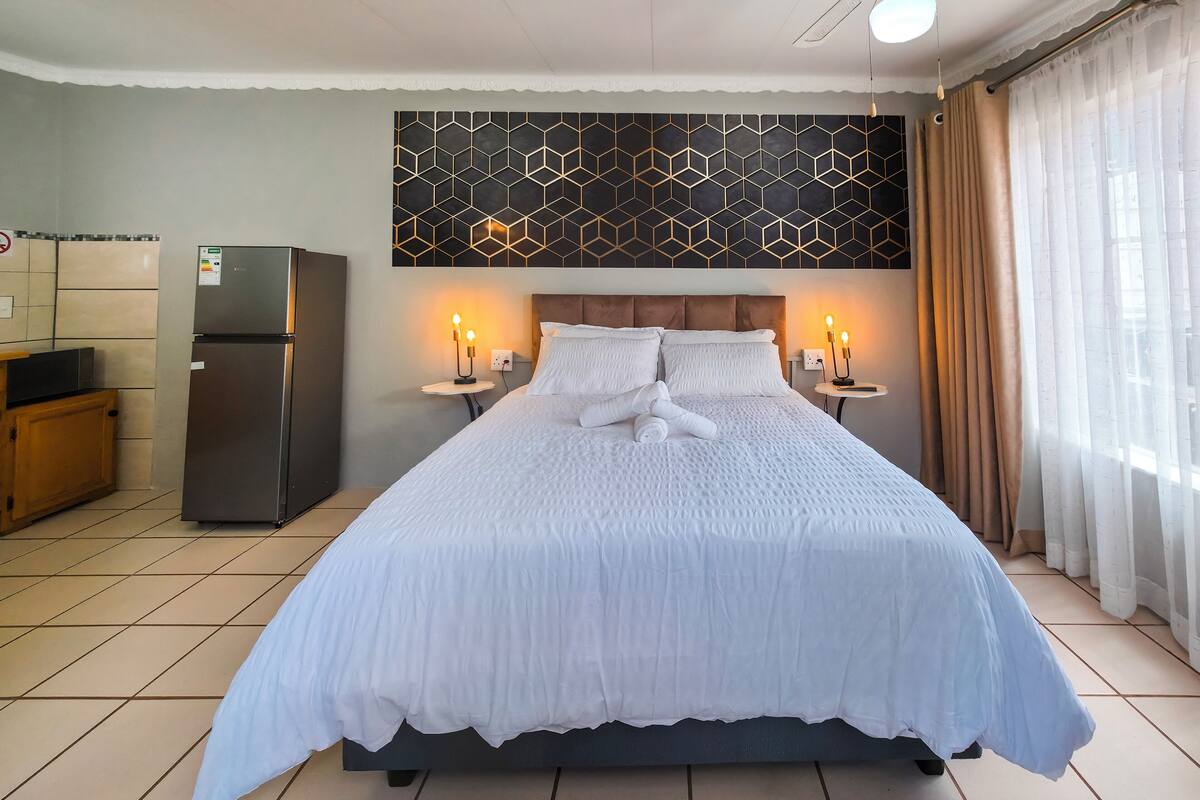 A hotel room with a large bed, a small kitchen area with a refrigerator, and a decorative wall panel.