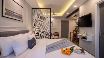A modern hotel room with a neatly made bed, a TV mounted on the wall, and a dining area visible through a partition.