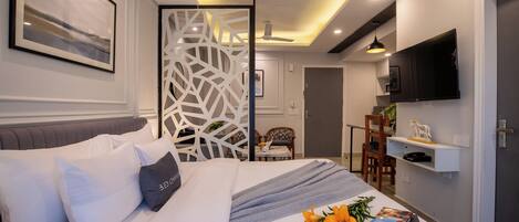 A modern hotel room with a neatly made bed, a TV mounted on the wall, and a dining area visible through a partition.