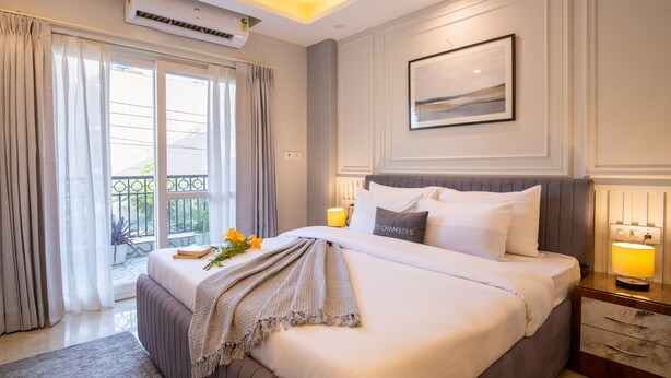 A well-lit bedroom with a large bed, a bedside table with a lamp, a framed picture on the wall, and a view of the outdoors through the window. - Bedchambers Apart hotel Sec 39 (Gurugram)