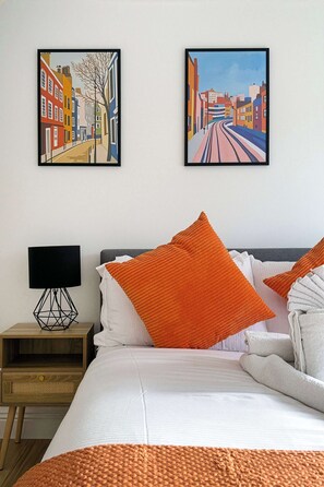 A hotel room with a bed, pillows, a nightstand, a lamp, and two framed artworks on the wall. - Spacious 2BR, 2 Bath with Balcony Sleeps 6 (Greater London)