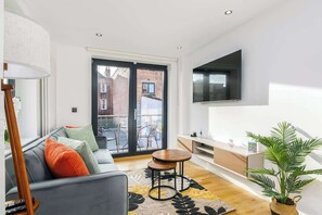 A modern living room with a sofa, a coffee table, a TV mounted on the wall, and a large window with a view of the outside.