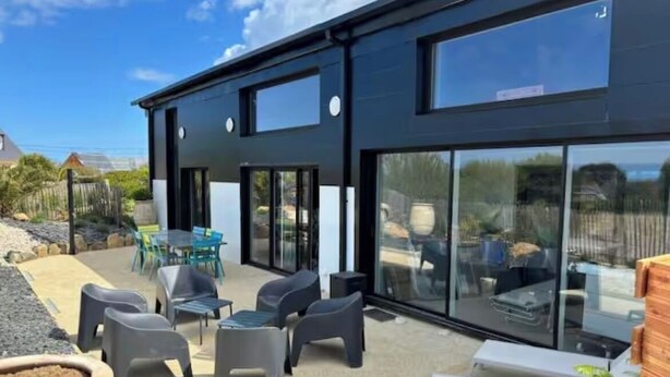 Un bâtiment moderne doté de grandes fenêtres en verre, d’un espace extérieur aménagé avec des chaises en plastique et d’une terrasse en béton.