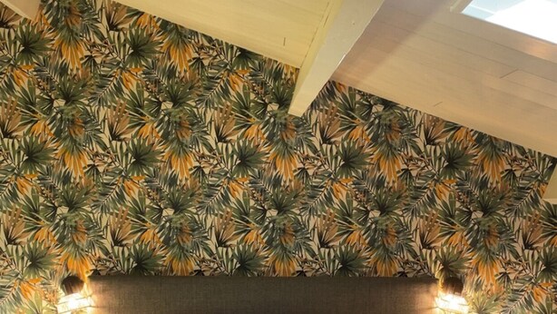 A neatly made bed with white linens and multiple pillows in a room with a tropical leaf-patterned wallpaper.
