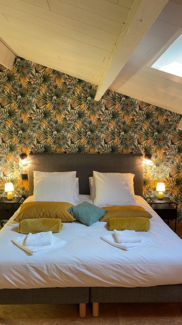 A neatly made bed with white linens and multiple pillows in a room with a tropical leaf-patterned wallpaper.