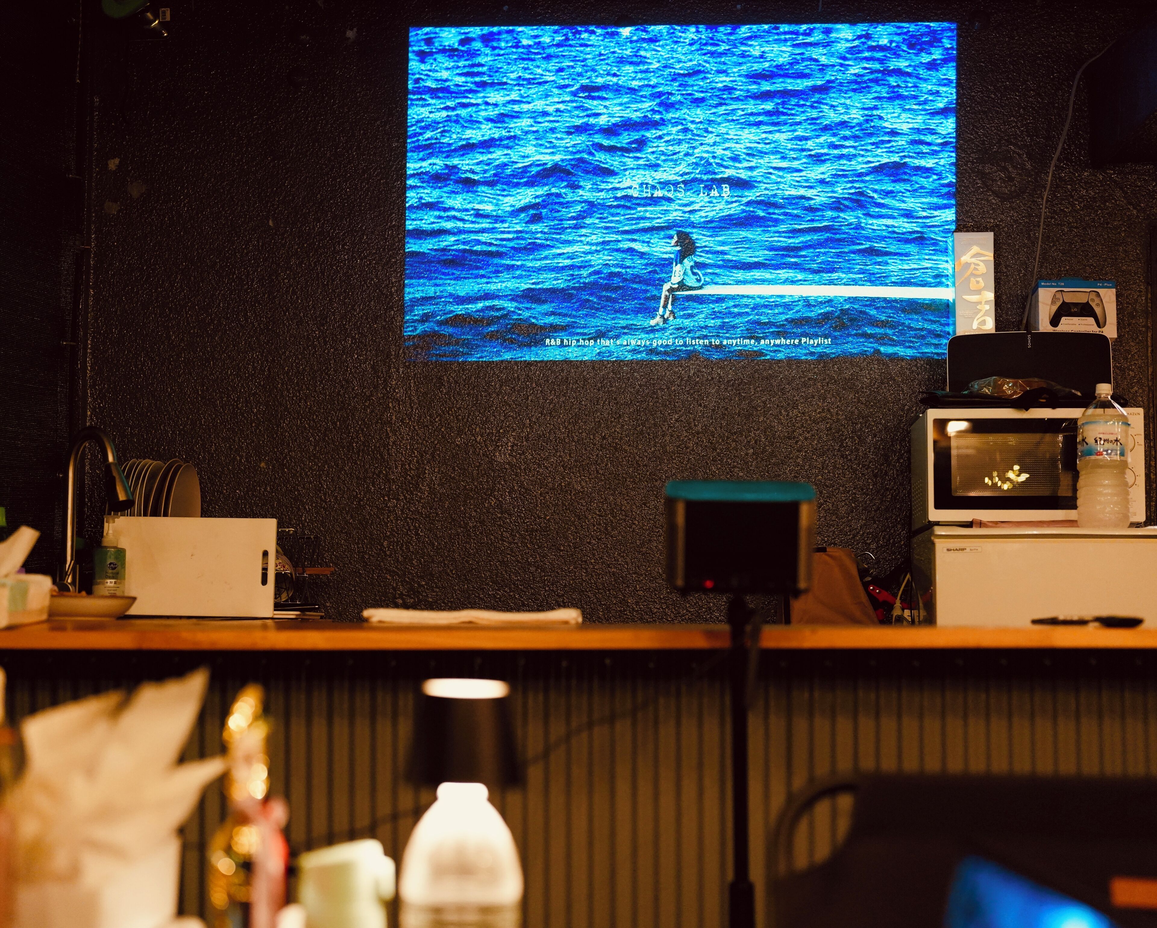 A room with a television displaying a blue ocean scene, a microwave, a water bottle, and a gaming console on a shelf.