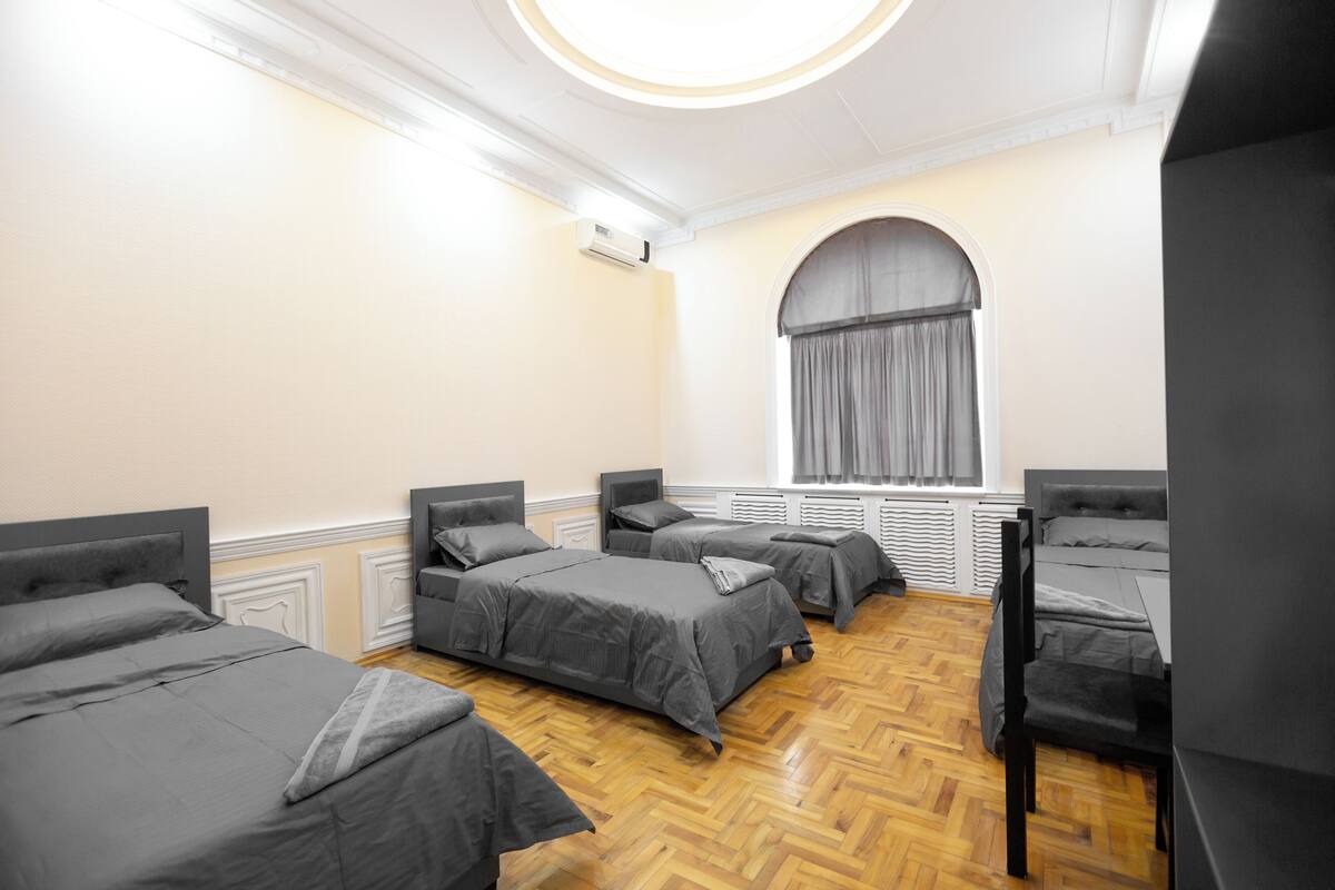 A hotel room with four beds arranged in a square layout, a large arched window with curtains, and a wooden parquet floor.