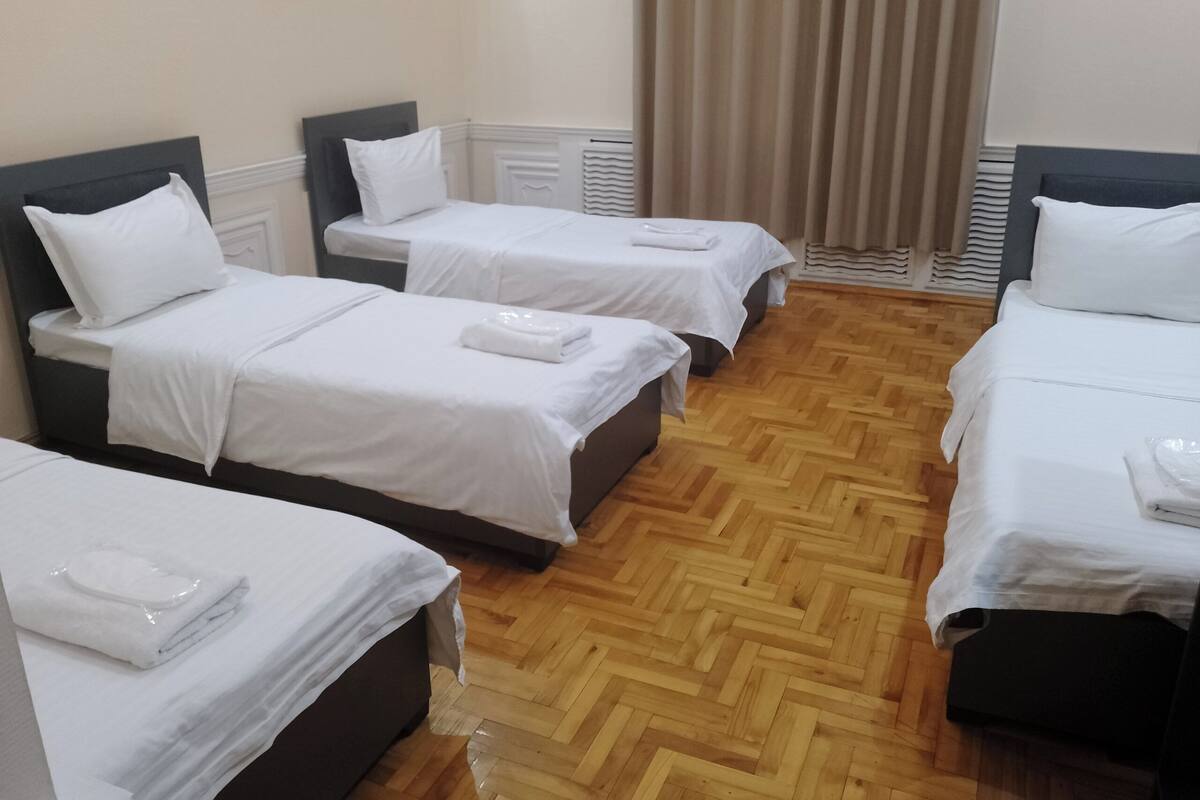 A hotel room with four single beds, wooden parquet flooring, and a window with curtains.