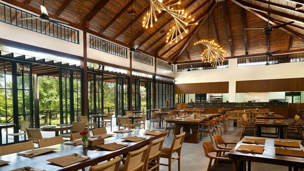 A spacious dining area with high ceilings and large windows, offering views of the outdoors. Tables are set with wicker chairs and centerpieces. - House Sira Lombok (Tanjung)