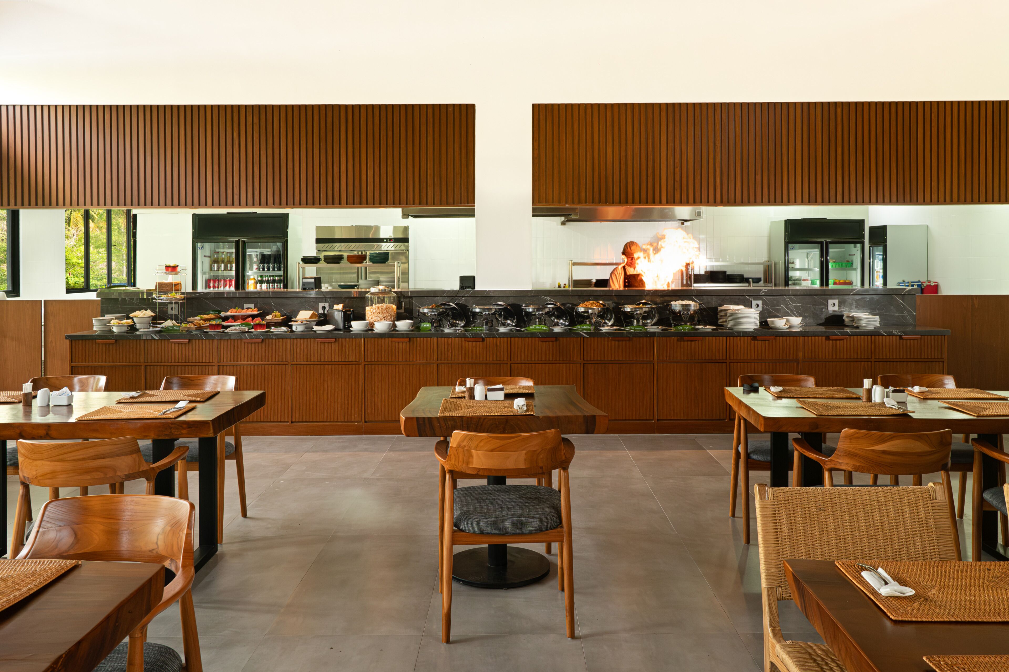 A modern restaurant with wooden paneling, a central kitchen with a chef, and multiple dining tables with chairs.