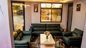 Een woonkamer met een groene zithoek, een glazen salontafel met een bloevas en grote ramen met uitzicht op de omgeving.