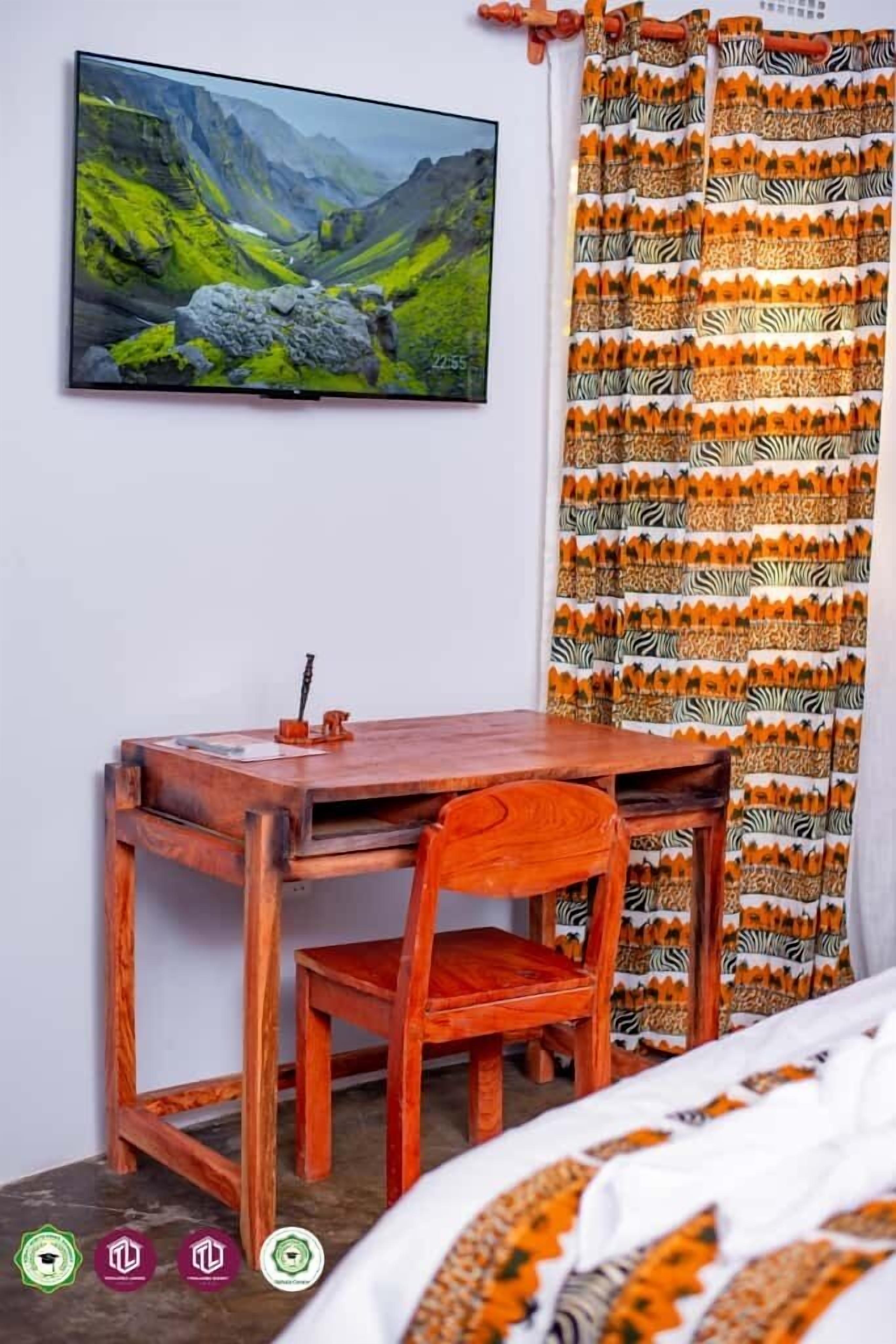 A room with a wooden desk, a chair, a bed with a patterned bedspread, a framed landscape painting, and zebra-striped curtains.