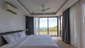 A bedroom with a large bed, an air conditioner, a ceiling fan, and a view of the ocean through a sliding glass door.