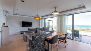 A modern kitchen with a marble island, a dining area with a wooden table and chairs, a built-in TV, and large windows offering a view of the ocean.