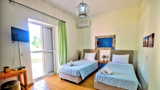 A room with two beds, a TV, a table, and a window with blinds. - Villazia Exclusive Estate (Corfu)
