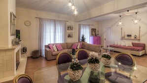 A modern living room with a beige sofa, a glass dining table, and purple chairs. There are decorative vases on the table, a pink cabinet, and a painting on the wall. - Villazia Exclusive Estate (Corfu)
