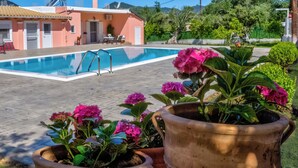 A swimming pool with a clear blue water surface, surrounded by potted plants with pink flowers, and a building with a tiled roof in the background. - Villazia Exclusive Estate (Corfu)