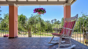 A covered patio with a rocking chair, a hanging flower basket, and a view of the garden. - Villazia Exclusive Estate (Corfu)