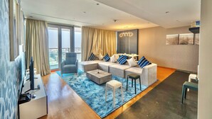 A modern living room with a sectional sofa, a coffee table, and a TV. There is a large window with curtains, a ceiling light, and a small table with a chair. - Luxury Vacation Home Nr Metro DWTC (Dubai)