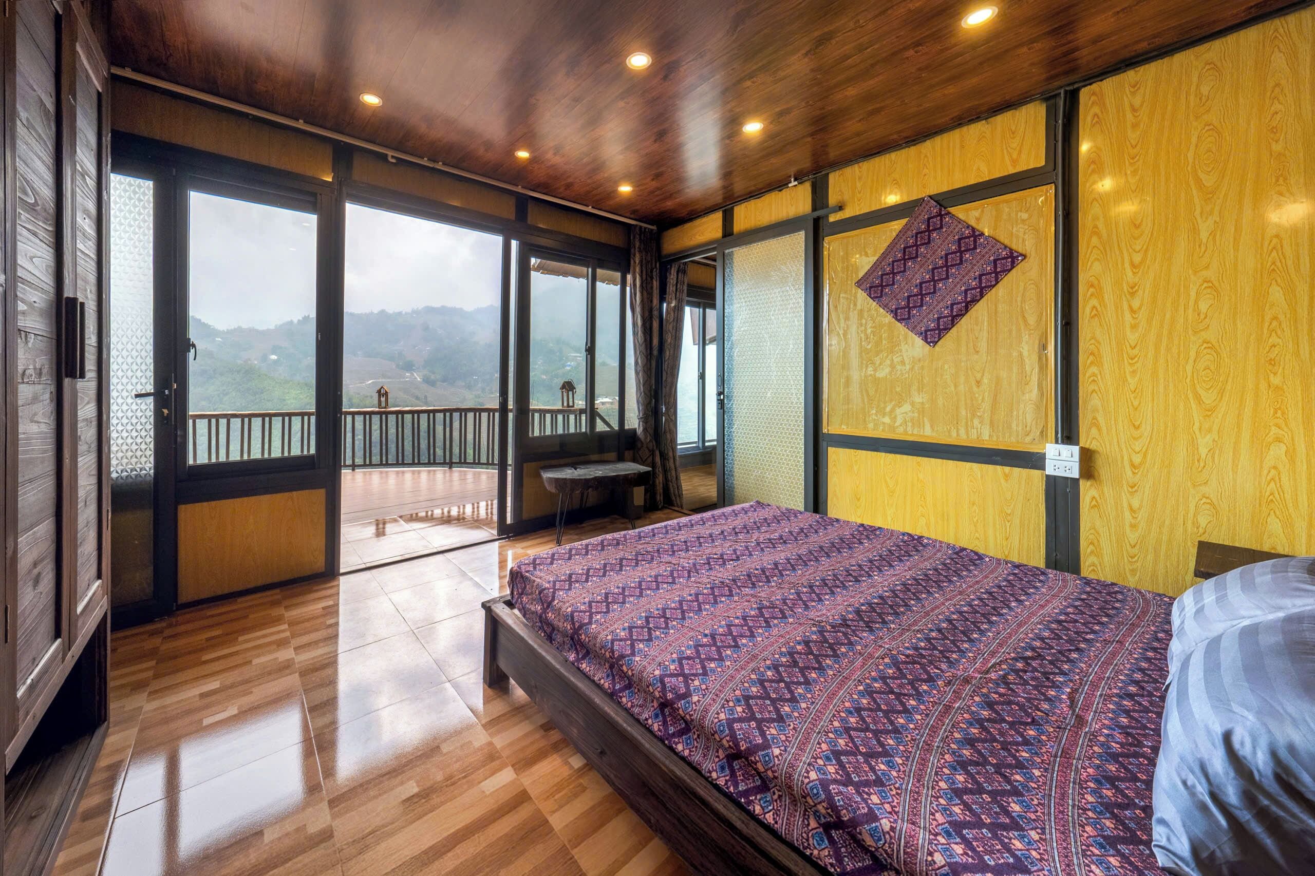 A bedroom with a large bed, wooden flooring, a balcony with a view, and a wooden ceiling.