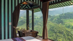 A bedroom with a large bed, wooden headboard, and a view of a green landscape through large windows. - Sapana Mountain Retreat (Sa Pa)