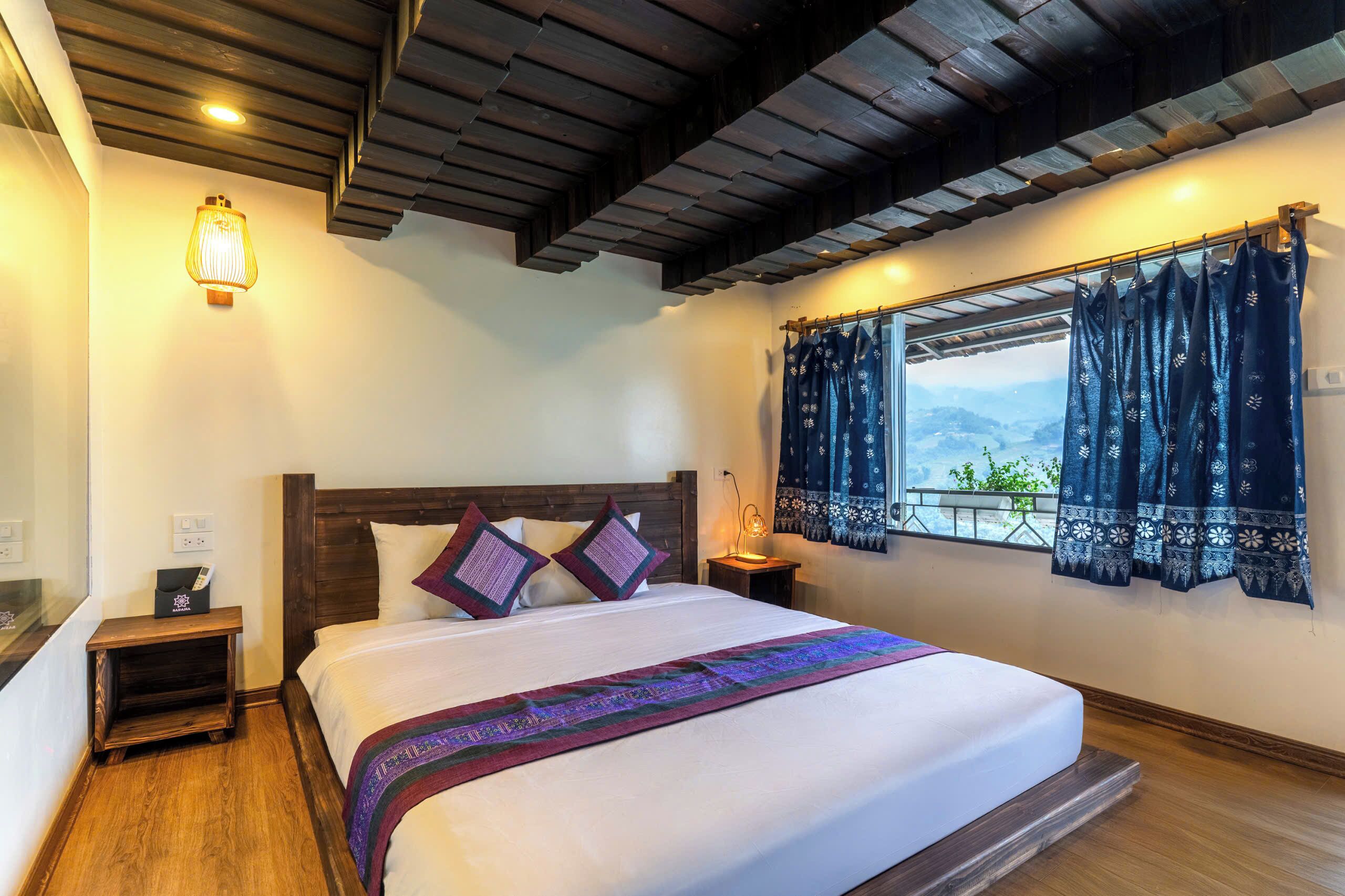 A bedroom with a wooden ceiling, a bed with a white bedspread, purple pillows, a nightstand with a lamp, and a window with patterned curtains.