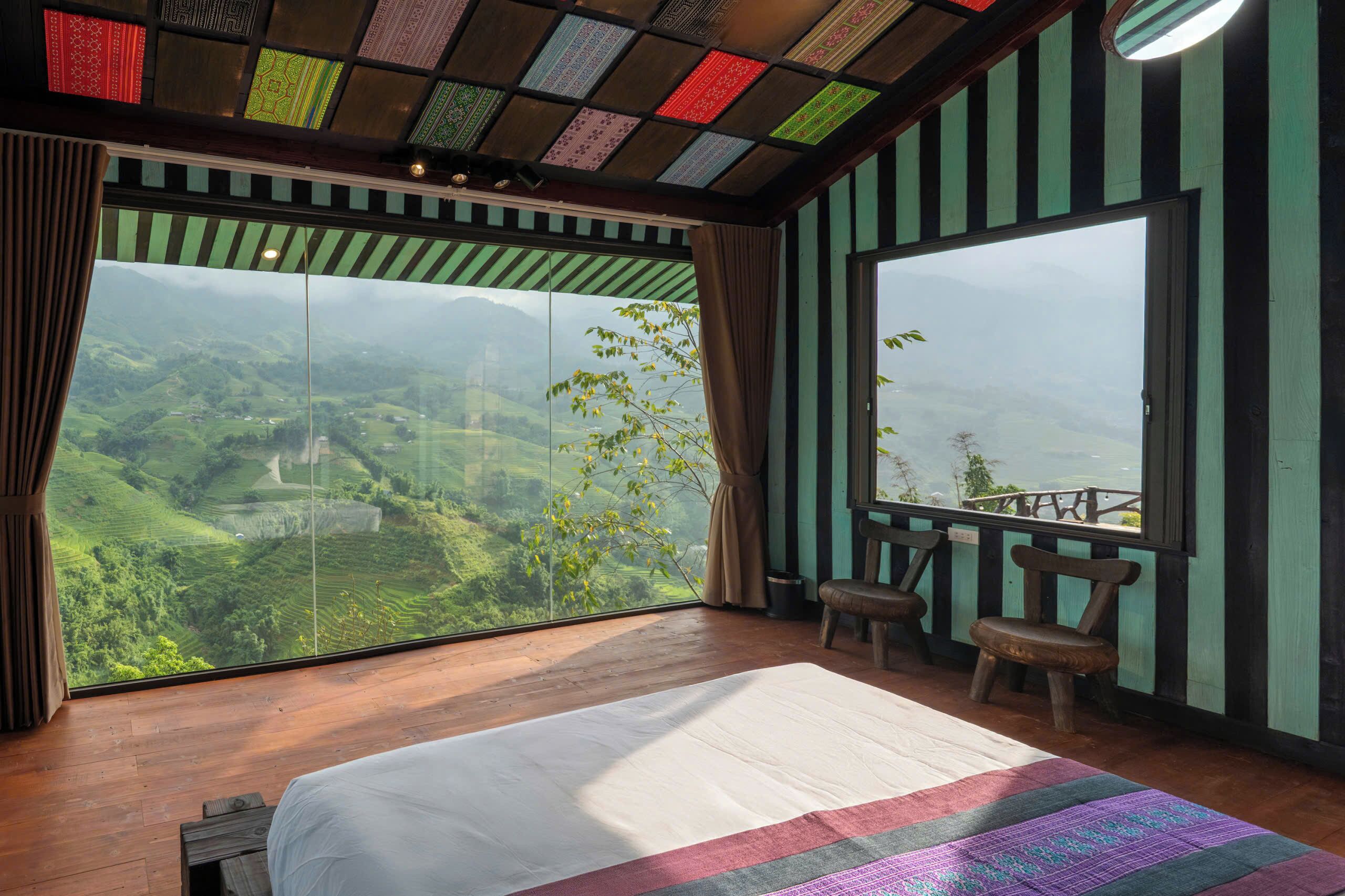A room with a large window offering a view of a green landscape. There is a bed with a white bedspread and a colorful blanket. Two wooden chairs are placed by the window.