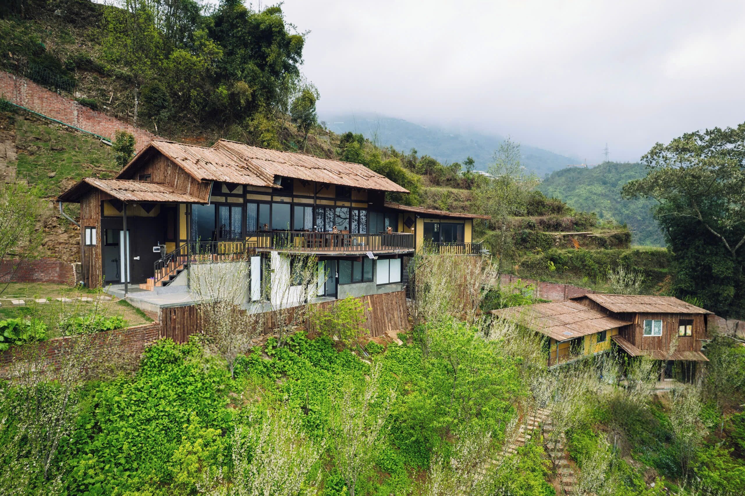Una casa moderna con un balcón grande, rodeada de abundante vegetación y colinas.