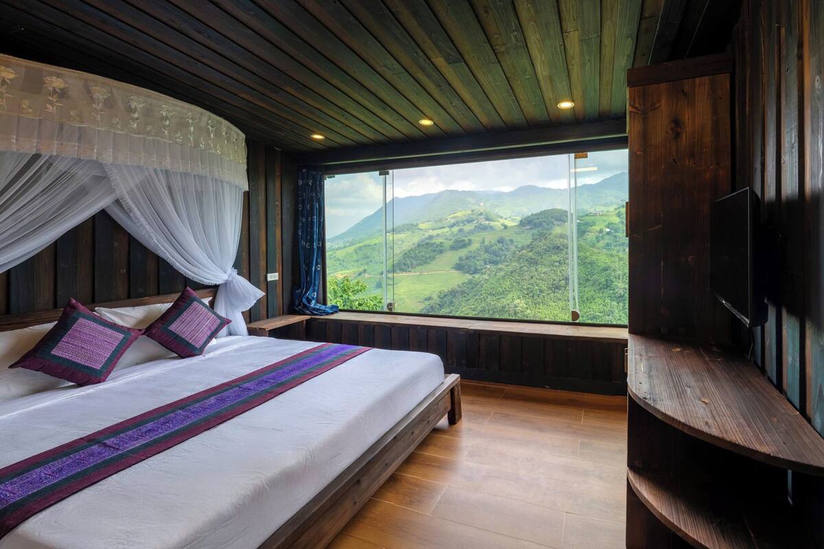 Een slaapkamer met een groot bed, een raam met uitzicht op glooiende heuvels, houten wanden en een tv op een houten dressoir.