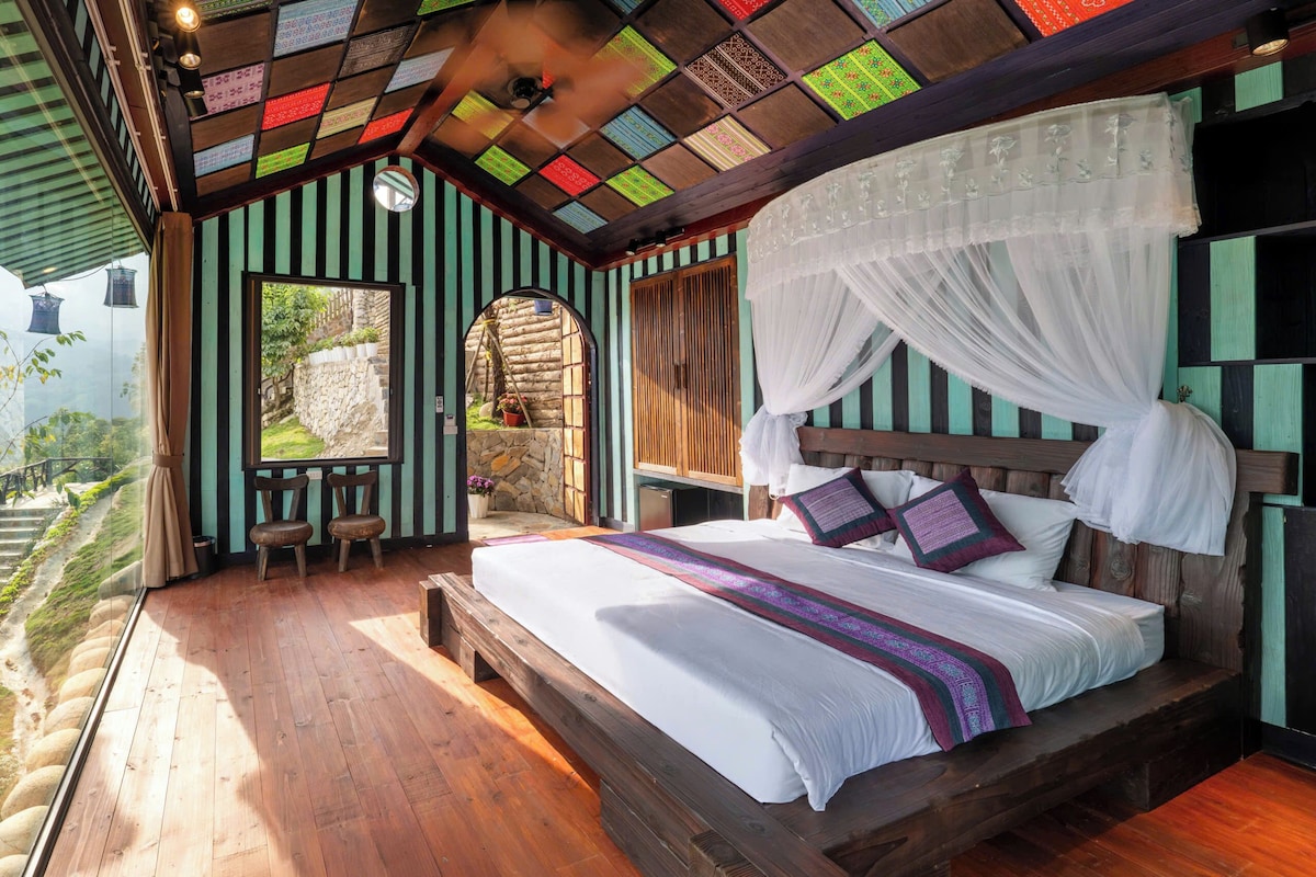 Una habitación con cama, ventana, banca de madera y pared de piedra.