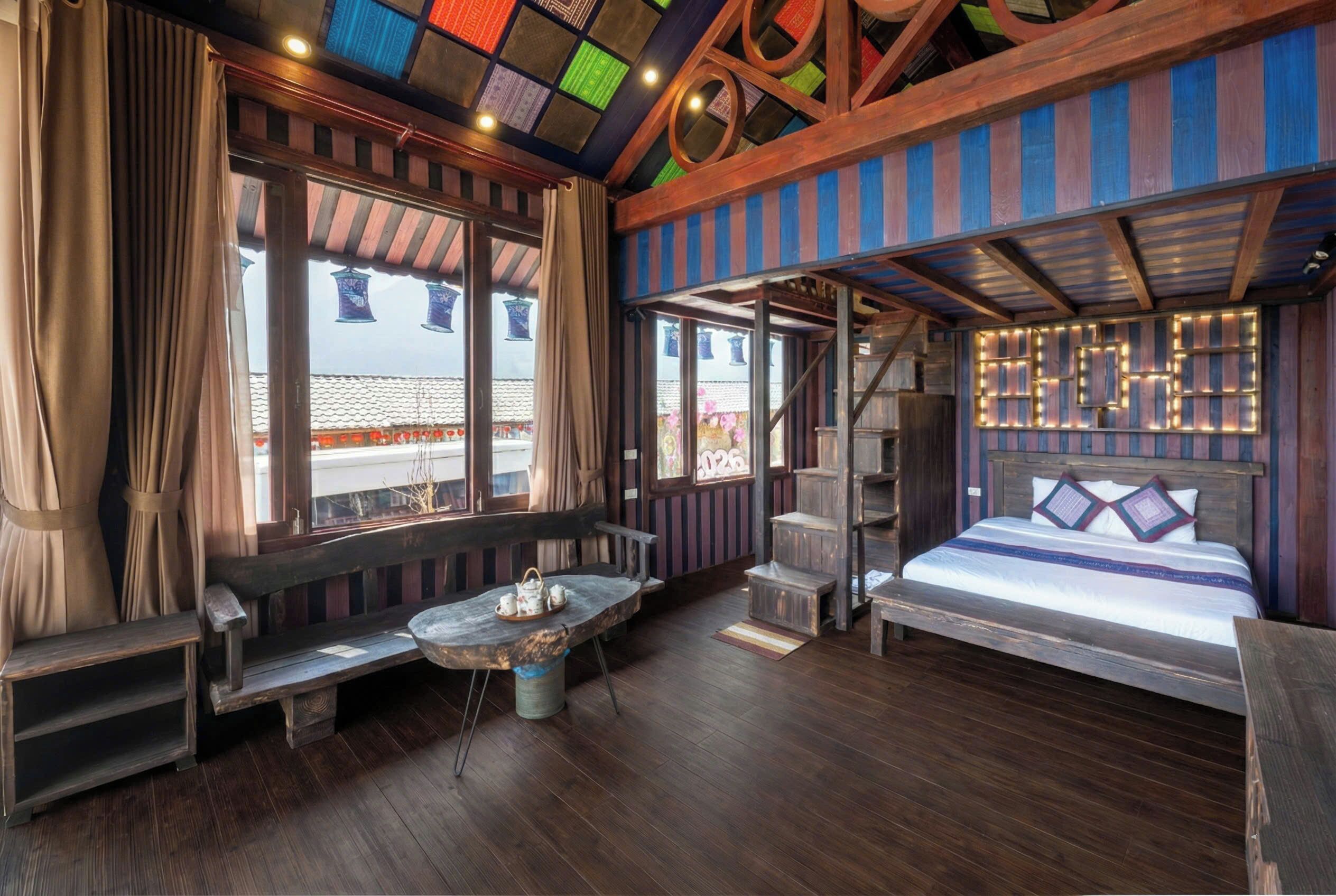 Una habitación con piso de madera, una cama con cabecera, un banco, una mesa con tetera y ventanas grandes con cortinas.
