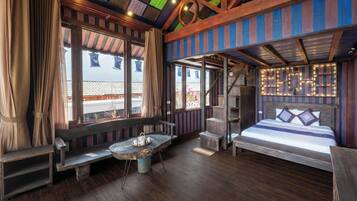Una habitación con piso de madera, una cama con cabecera, un banco, una mesa con tetera y ventanas grandes con cortinas.