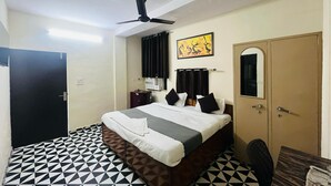 A hotel room with a bed, a ceiling fan, a wall-mounted air conditioner, a door, a small table, and a chair. - Roomshala Hotel South Embassy (New Delhi)