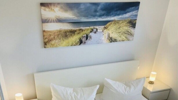 A neatly made bed with white linens and pillows, two bedside tables with lamps, and a painting of a coastal landscape above the bed.