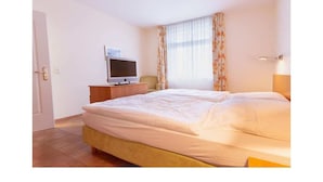 A hotel room with a bed, a television on a stand, a window with curtains, and a door. - Suurhusen - Greetsiel (Greetsiel)