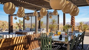 An outdoor dining area with wicker hanging lamps, cushioned seating, and a view of the desert landscape. - Iconic 6BR Desert Retreat w/ Pool, Pickleball & Fire Pit Joshua Tree Mansion by Avantstay (Joshua Tree)